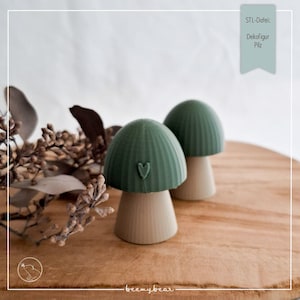 May include: Two decorative mushroom figurines. The mushroom caps are a muted green with a heart detail, and the stems are a light beige. The mushrooms are displayed on a wooden surface. The text "STL-Datei: Dekofigur Pilz" is in the upper right corner.