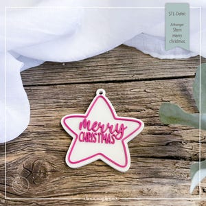 May include: A white star-shaped ornament with a pink outline and the words "Merry Christmas" in pink script. The ornament has a small hole at the top for hanging. The background is a wooden surface.