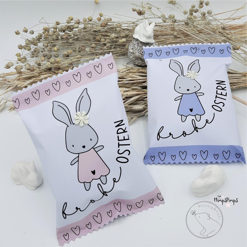 以下が含まれることがあります： ウサギのイラストと「Frohe Ostern」（ハッピーイースター）の文字が書かれた白いギフトバッグが2つ。左側のバッグにはピンクのウサギと、縁にピンクのハートが描かれている。右側のバッグには青いウサギと、縁に青いハートが描かれている。