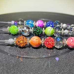 Peut inclure: Trois ensembles de boucles d'oreilles perlées avec des perles décoratives colorées. Les boucles d'oreilles présentent une variété de styles de perles, dont des motifs en cristal, à paillettes et marbrés. Les couleurs incluent l'orange, le rouge, le rose, le vert, le bleu et le violet.