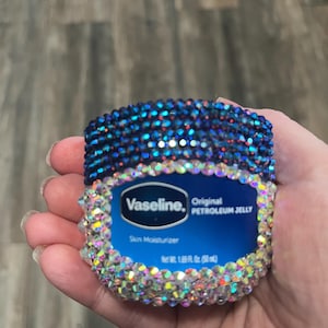 Puede incluir: Un tarro azul y blanco de Vaseline Original Petroleum Jelly adornado con pedrería. El tarro está cubierto de pedrería iridiscente y tiene el texto "Vaseline. Original Petroleum Jelly. Skin Moisturizer. Net Wt. 1.75 oz (50 g)"