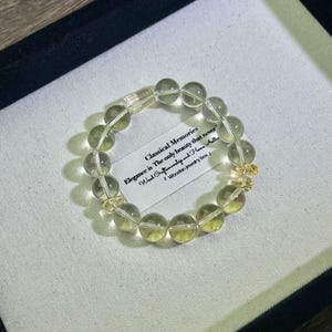 May include: A bracelet made of translucent, round, light-yellow beads. The bracelet is displayed in a black box with a white interior. A small tag with the text "Classical Memories" is visible.