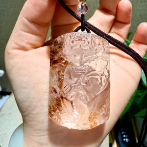 May include: A rectangular clear quartz pendant featuring an intricate relief carving of a mythical figure with large eyes and ornate headwear. The stone displays natural reddish-brown inclusions and hangs from a dark brown cord with a small bead.