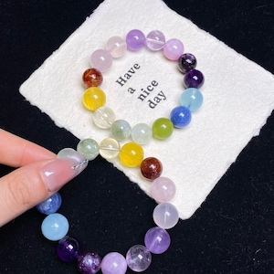 May include: A multi-colored beaded bracelet with various shades of purple, pink, yellow, green, and blue gemstones. The bracelet is resting on a white cloth with the text "Have a nice day".