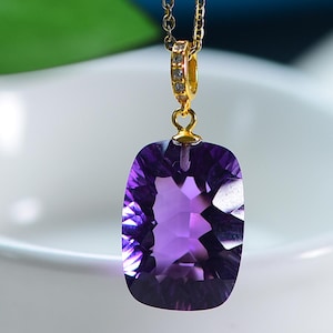 Op de afbeelding: Een rechthoekige, gefacetteerde amethist hanger hangt aan een delicate gouden ketting. De dieppaarse edelsteen is gezet in een gouden bezel en de ketting heeft een kleine, decoratieve gouden ring met ingebedde heldere stenen.