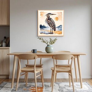 K&ouml;nnte beinhalten: Ein gerahmter Aquarelldruck eines Reihers, der an einem Strand steht, mit Sonne und Landschaft im Hintergrund. Der Reiher ist in Grau-, Braun- und Blaut&ouml;nen dargestellt. Der Rahmen hat eine helle Holzfarbe. Der Druck wird in einem Esszimmer ausgestellt.