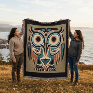 May include: A woven blanket featuring a stylized owl design in shades of beige, teal, brown, and black. The owl has large, circular eyes and a symmetrical pattern. The blanket is held up against a backdrop of the ocean and sky.
