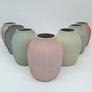 Könnte beinhalten: Eine Sammlung von acht dekorativen Vasen in verschiedenen Farben, darunter Rosa, Grün, Grau und Beige. Jede Vase hat eine gerippte Textur und eine breite Öffnung. Die Vasen sind in einer Gruppe angeordnet und zeigen ihre verschiedenen Farbtöne.