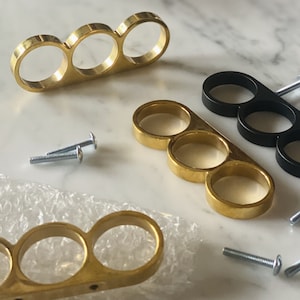 May include: Three gold-toned metal rings with three holes each, arranged in a row. The rings are connected to each other. The rings are on a white surface.