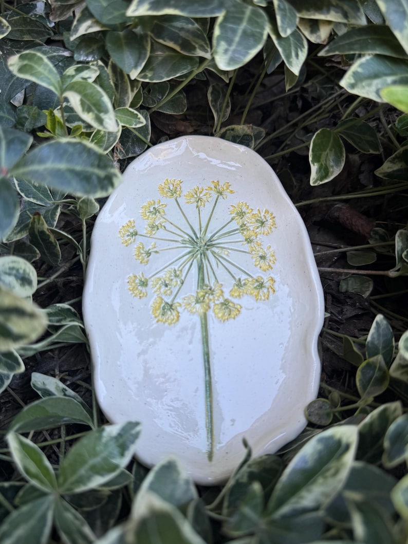 May include: A cream-colored, oval-shaped ceramic dish featuring a detailed illustration of a yellow flower with a green stem. The dish is surrounded by green and white foliage, creating a natural, garden-inspired aesthetic.