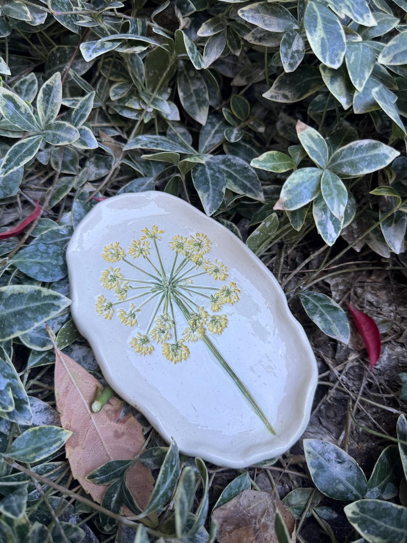 May include: A handmade, off-white ceramic dish with a floral design. The dish features a yellow and green botanical illustration of a flower. The oval-shaped dish is surrounded by green and white foliage.