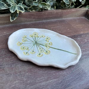 May include: A handmade, off-white ceramic dish with a floral design. The dish has an irregular, organic shape and features a green stem and yellow flower design. The dish is on a wooden surface.