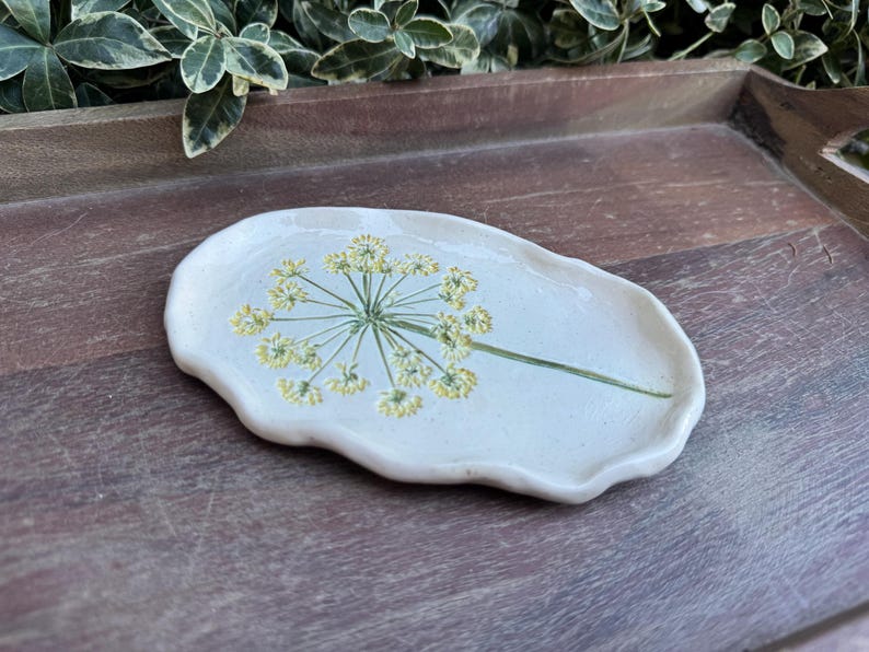 May include: A handmade, off-white ceramic dish with a floral design. The dish has an irregular, organic shape and features a green stem and yellow flower design. The dish is on a wooden surface.