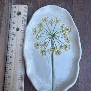 May include: Off-white ceramic serving dish with a raised floral design. The dish is oval-shaped and features a green stem with yellow flower clusters. The dish measures approximately 10 inches long.