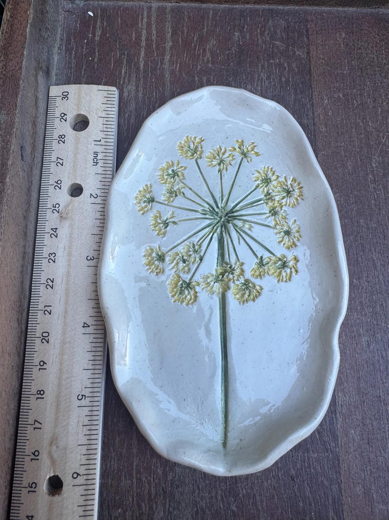 May include: Off-white ceramic serving dish with a raised floral design. The dish is oval-shaped and features a green stem with yellow flower clusters. The dish measures approximately 10 inches long.
