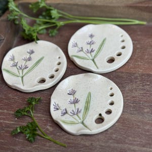 May include: Three off-white ceramic dishes with a lavender sprig design. Each dish has a series of small holes along the edge. The lavender is painted in purple and green. The dishes are on a wooden tray.