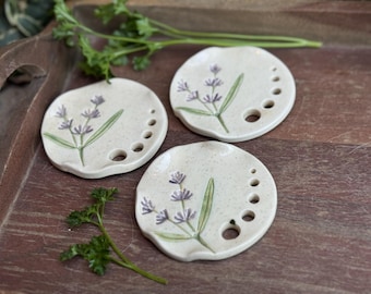 May include: Three off-white ceramic dishes with a lavender sprig design. Each dish has a series of small holes along the edge. The lavender is painted in purple and green. The dishes are on a wooden tray.