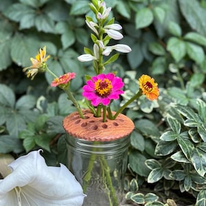 May include: A clear glass jar with a brown ceramic flower frog holding colorful flowers. The flowers include a bright pink zinnia, yellow and orange blooms, and white flowers. The arrangement is set against a backdrop of green foliage.
