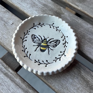 May include: A small, round, off-white ceramic dish with a scalloped edge. The dish features a hand-painted bee with yellow and black stripes, surrounded by a black floral wreath design. The dish is approximately 4 inches in diameter.