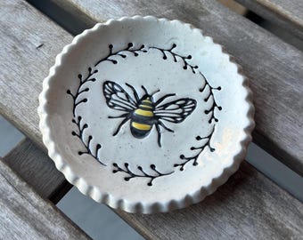 May include: A small, round, off-white ceramic dish with a scalloped edge. The dish features a hand-painted bee with yellow and black stripes, surrounded by a black floral wreath design. The dish is approximately 4 inches in diameter.
