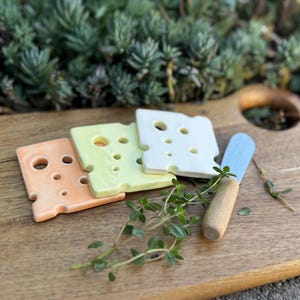 May include: Three ceramic cheese-shaped serving plates in orange, yellow, and white, with a wooden cutting board and a blue and wooden butter knife.