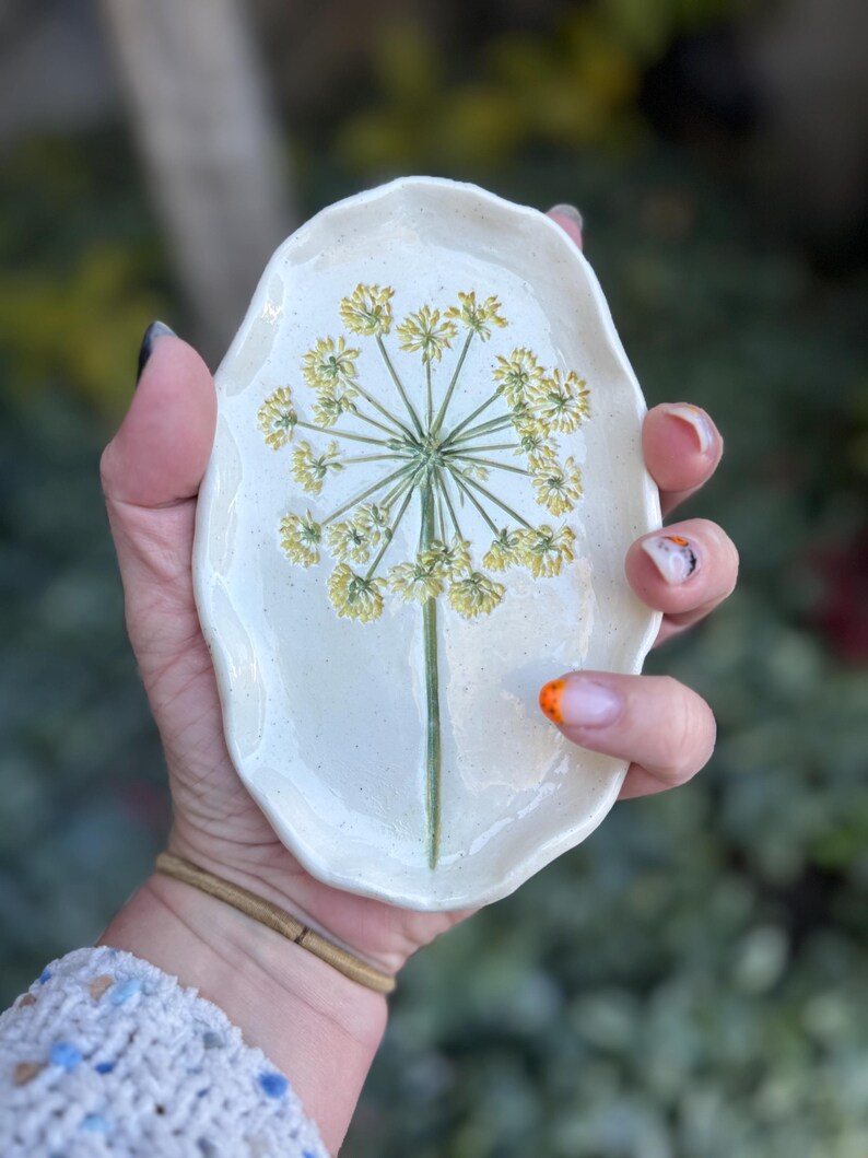 May include: A small, off-white ceramic dish with an organic shape. The dish features a hand-painted yellow and green floral design in the center. The dish is held in a person's hand, with a blurred green background.