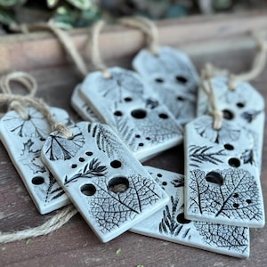 May include: A set of six white ceramic gift tags with black leaf designs and holes for string. The tags are tied together with twine.