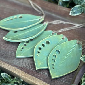 May include: A set of five leaf-shaped ceramic ornaments in a glossy green glaze. Each piece features a central vein detail and a series of five circular cutouts of varying sizes along one side. Each ornament is attached to a thin jute twine string.