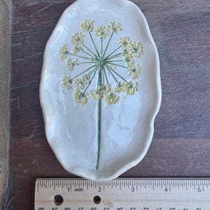 May include: A handmade, off-white ceramic dish with a floral design. The dish is approximately 5 inches long. The floral design features a green stem and yellow flower clusters. The dish has a unique, organic shape and is placed on a wooden surface.