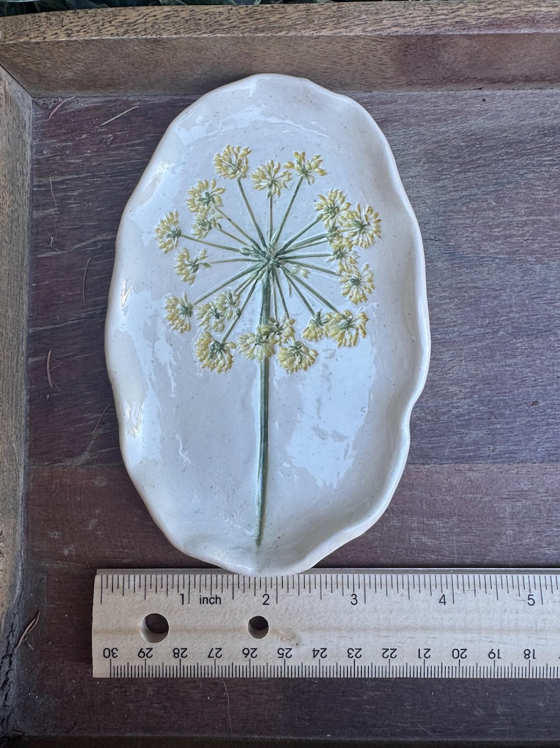 May include: A handmade, off-white ceramic dish with a floral design. The dish is approximately 5 inches long. The floral design features a green stem and yellow flower clusters. The dish has a unique, organic shape and is placed on a wooden surface.