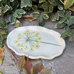 May include: A handmade, light blue ceramic serving dish with a floral design. The dish has a unique, organic shape and features a detailed illustration of a yellow and green flower. The dish is approximately 10 inches long.
