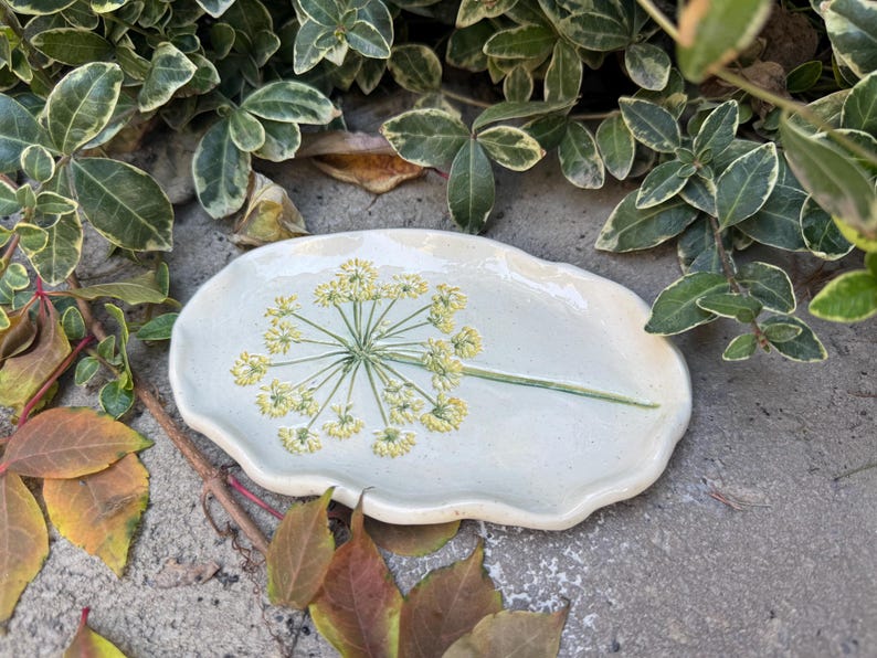 May include: A handmade, light blue ceramic serving dish with a floral design. The dish has a unique, organic shape and features a detailed illustration of a yellow and green flower. The dish is approximately 10 inches long.