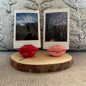 May include: Two Polaroid style photo frames with red and pink lips as bases, sitting on a wooden slice.