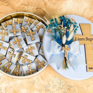 Puede incluir: Una lata redonda abierta llena de chocolates envueltos, cada uno adornado con una cruz de madera. La tapa presenta una cruz de madera, el texto "Liam Baptism", una fecha, un ramo y un lazo.