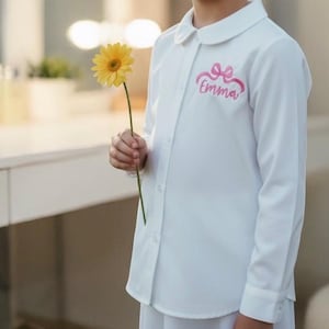 May include: A white long-sleeved shirt with a rounded collar and button-down front. The shirt features a pink bow and the name "Emma" embroidered on the chest. A yellow gerbera daisy is held in the hand.
