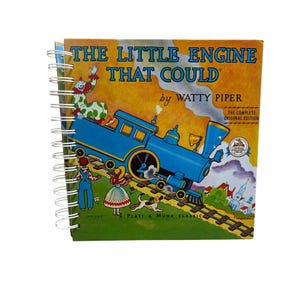 May include: A spiral-bound book with the title "THE LITTLE ENGINE THAT COULD" by Watty Piper. The cover features a blue cartoon train on tracks, with illustrations of a clown, children, and a dog. The background is yellow and green.
