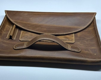 May include: A brown leather laptop sleeve with a top flap and a handle. The sleeve has a pocket on the front with the text "MAJAD Med" embossed on it.