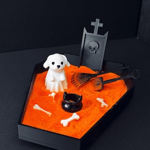May include: A miniature Halloween-themed diorama in a black coffin-shaped box. It features a white ghost dog, a tombstone with a skull, orange sand, bones, a cauldron, and miniature gardening tools.
