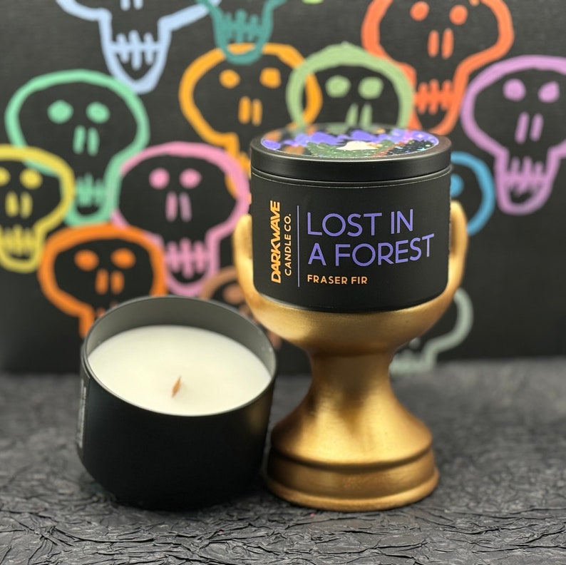May include: A black candle jar with a gold lid and a gold stand. The candle is labelled "Darkwave Candle Co. Lost in a Forest Fraser Fir". The candle is surrounded by colourful skull drawings.
