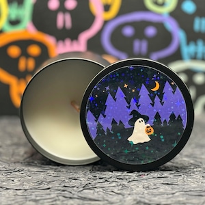 May include: A black candle jar with a white candle inside. The lid of the jar has a colourful design of a ghost holding a pumpkin in a forest at night.