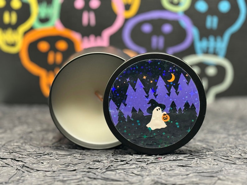 May include: A black candle jar with a white candle inside. The lid of the jar has a colourful design of a ghost holding a pumpkin in a forest at night.