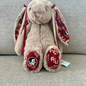 May include: A plush bunny rabbit in a seated position. The bunny is light brown with long ears lined with a maroon and gold pattern. The bunny's feet also feature the maroon and gold pattern with the letters "FSU" and a Seminole head logo. Red and cream ribbons are tied to the ears.