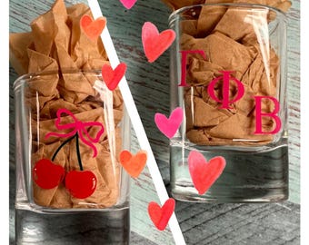May include: Two clear shot glasses with pink designs. One glass has a pink cherry design with a pink bow. The other glass has the letters "ΓΦΒ" in pink.