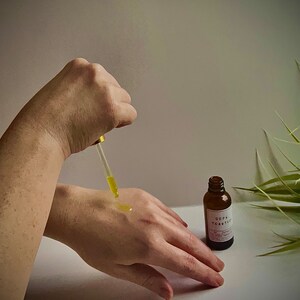 May include: A person's hand applying a clear liquid from a dropper bottle to their other hand. The bottle is labeled "Oop Tea Tree" and is sitting on a white surface. A green plant is in the background.
