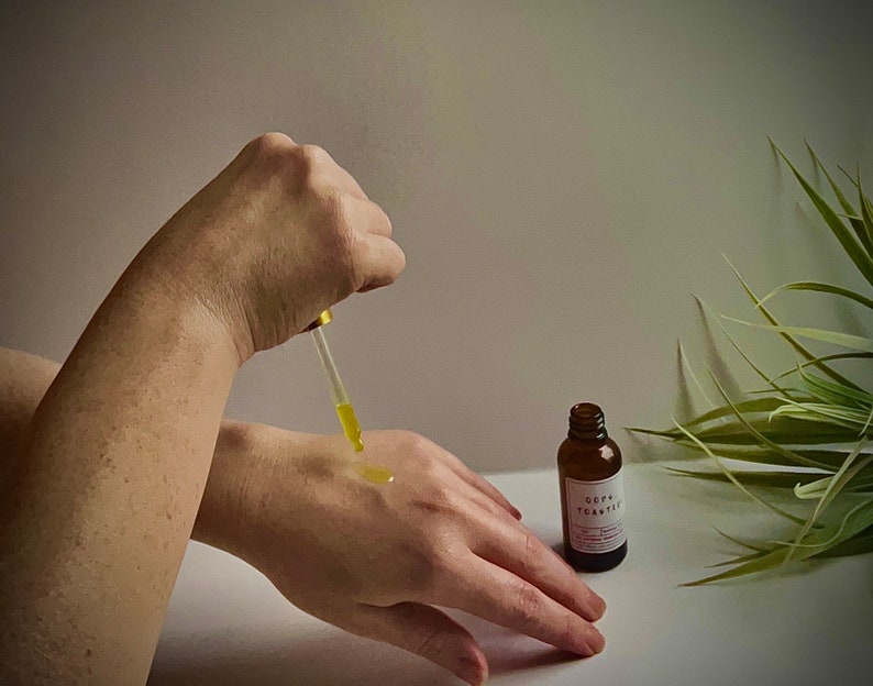 May include: A person's hand applying a clear liquid from a dropper bottle to their other hand. The bottle is labeled "Oop Tea Tree" and is sitting on a white surface. A green plant is in the background.