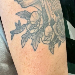 May include: A black and grey tattoo of a Native American wearing a headdress with feathers, flowers, and a hummingbird. The tattoo is on a person's upper arm. The text "The Next Day" is visible below the tattoo.
