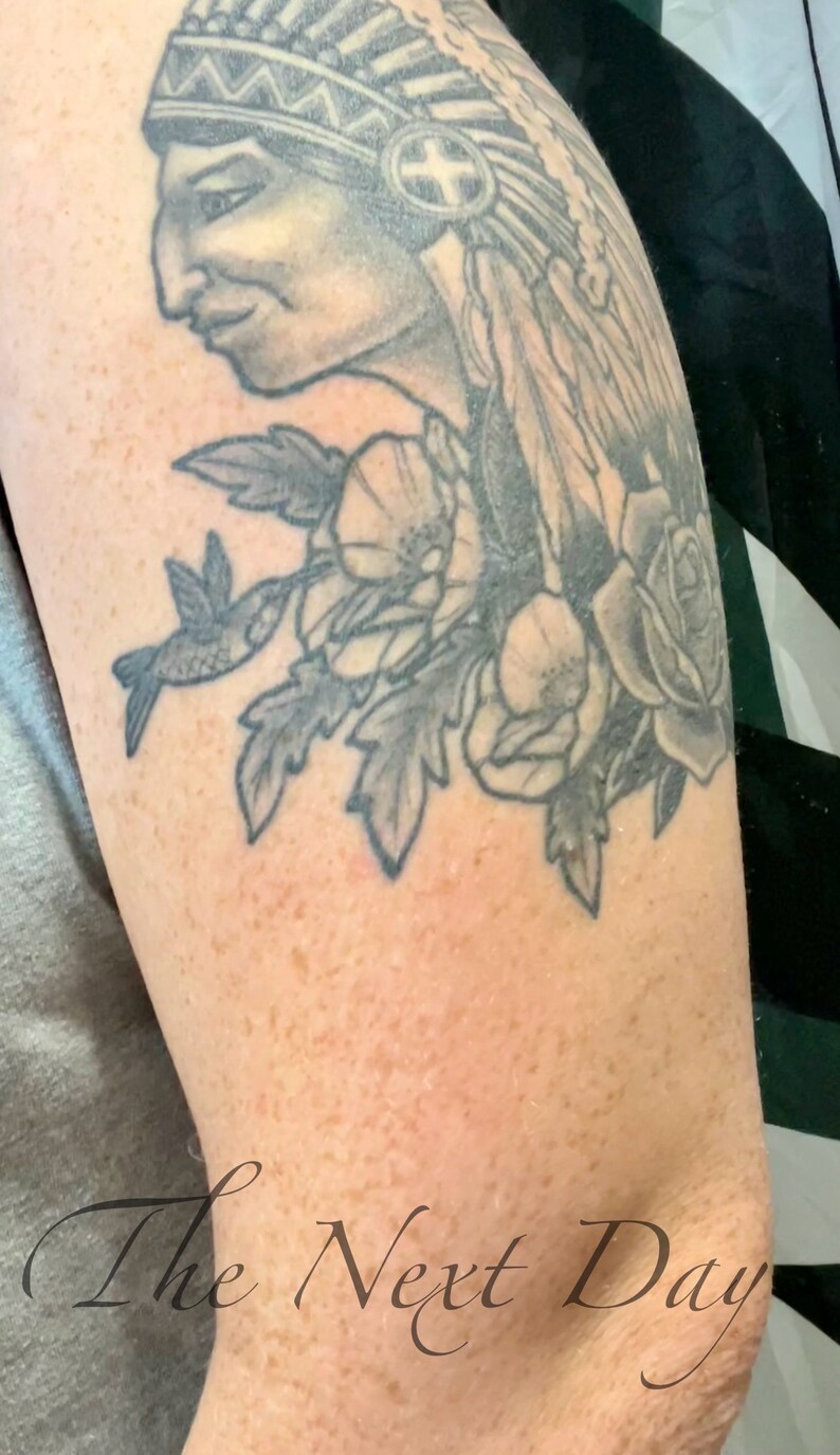 May include: A black and grey tattoo of a Native American wearing a headdress with feathers, flowers, and a hummingbird. The tattoo is on a person's upper arm. The text "The Next Day" is visible below the tattoo.