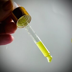 May include: A glass dropper with a gold top filled with a yellow liquid. The liquid is dripping from the end of the dropper.