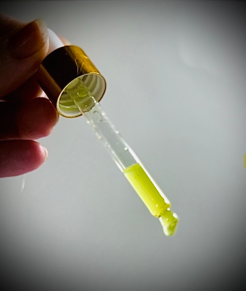May include: A glass dropper with a gold top filled with a yellow liquid. The liquid is dripping from the end of the dropper.