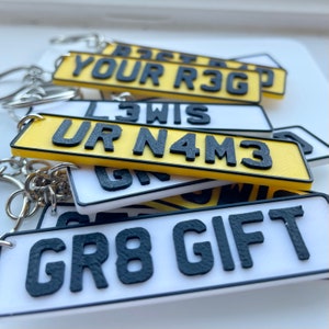 May include: A set of six keychains featuring personalised number plates. The plates are white with black lettering and a yellow border. The plates feature the text "YOUR R3G", "UR N4M3", "GR8 GIFT", and other personalised names.